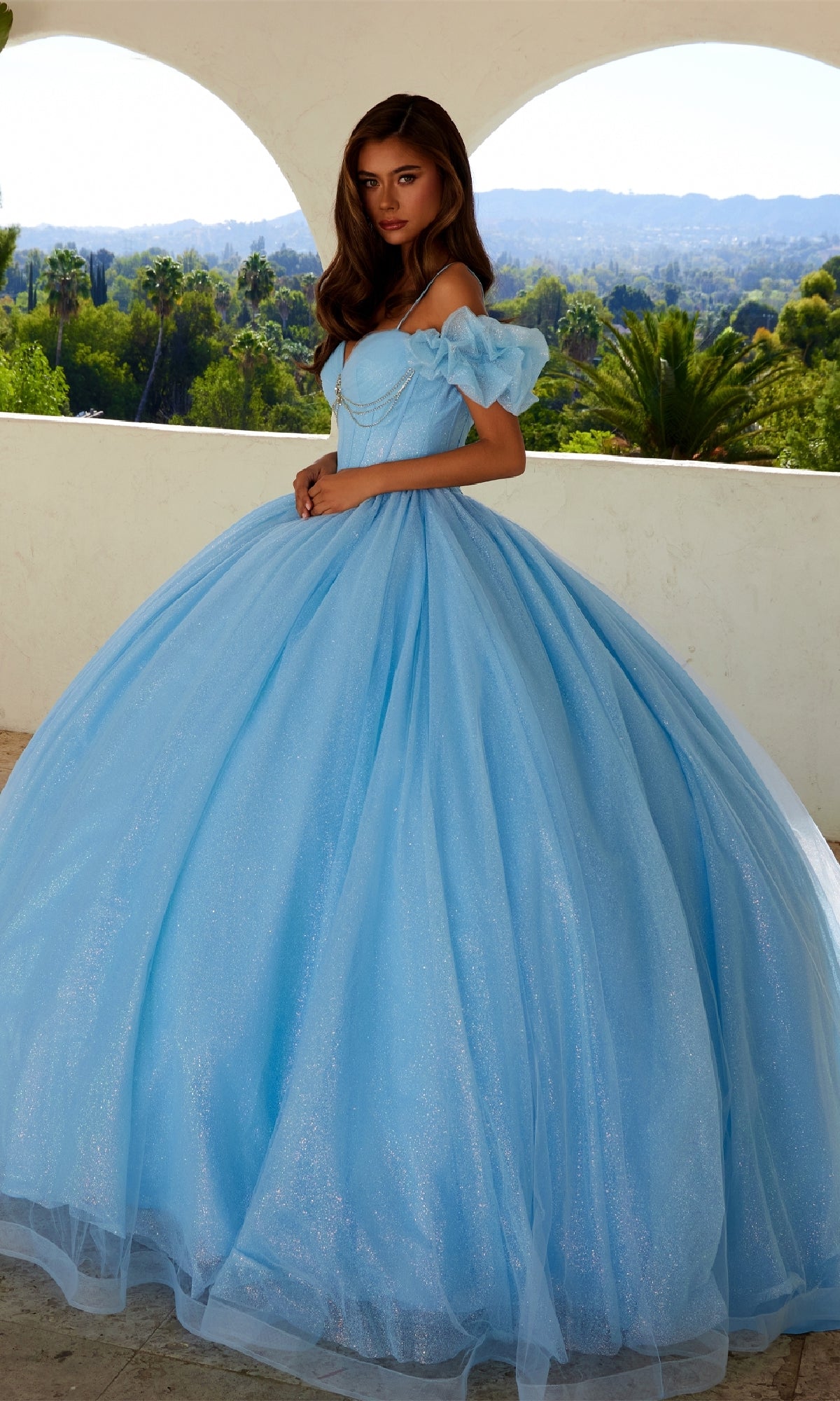 A woman stands outdoors in the Quinceanera Dress JT1487H By Juliet, a voluminous light blue gown with puff off-the-shoulder sleeves. Green trees and distant hills enhance the enchanting scene.